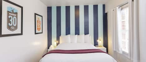 A neatly arranged hotel room with a large bed, striped wall, framed pictures, and a window with curtains.