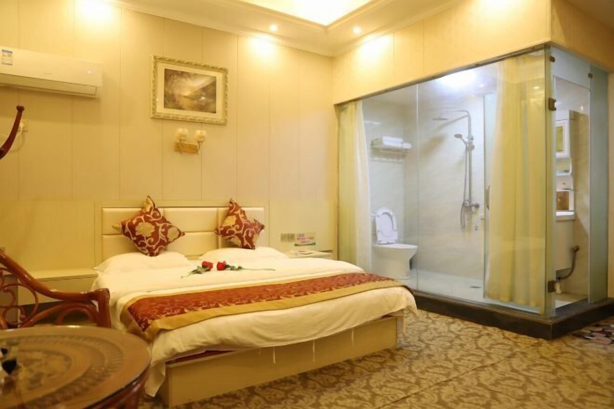 A hotel room with a bed, a glass-enclosed shower, a bedside table, and a chair.