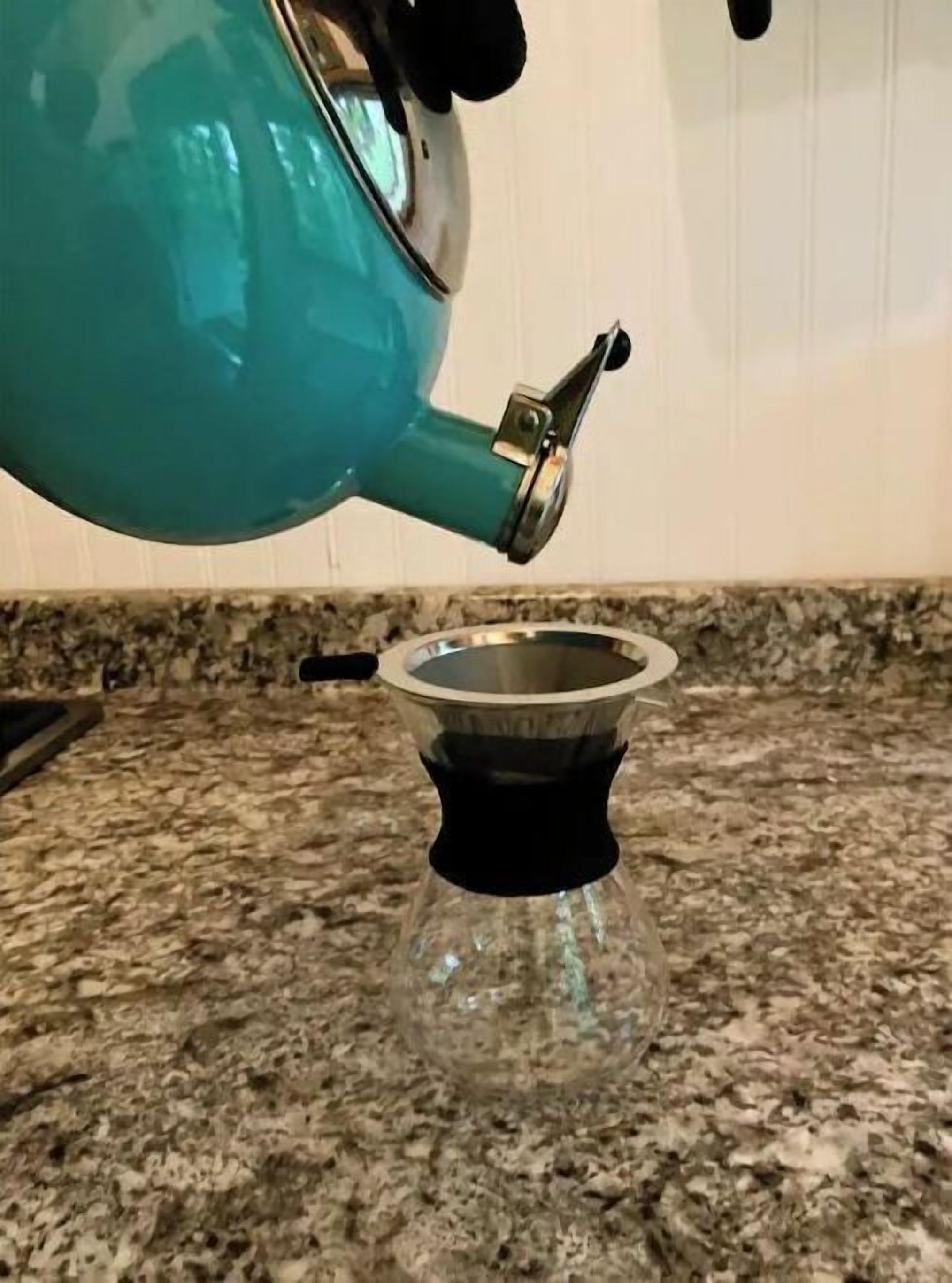 Una tetera color turquesa con asa negra y pico metálico vertiendo en una jarra de vidrio sobre una encimera de granito.