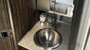 A compact, modern bathroom with a built-in sink and mirror, a small storage cabinet, and a bunk bed visible through the mirror. - Zion's Cozy Airstream #2 (Apple Valley)