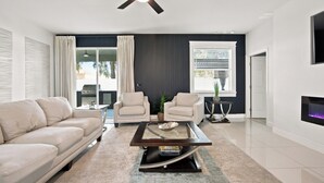 Una sala moderna con un sofá, dos sillones, una mesa de centro, un ventilador de techo, una chimenea y un ventanal con persianas.