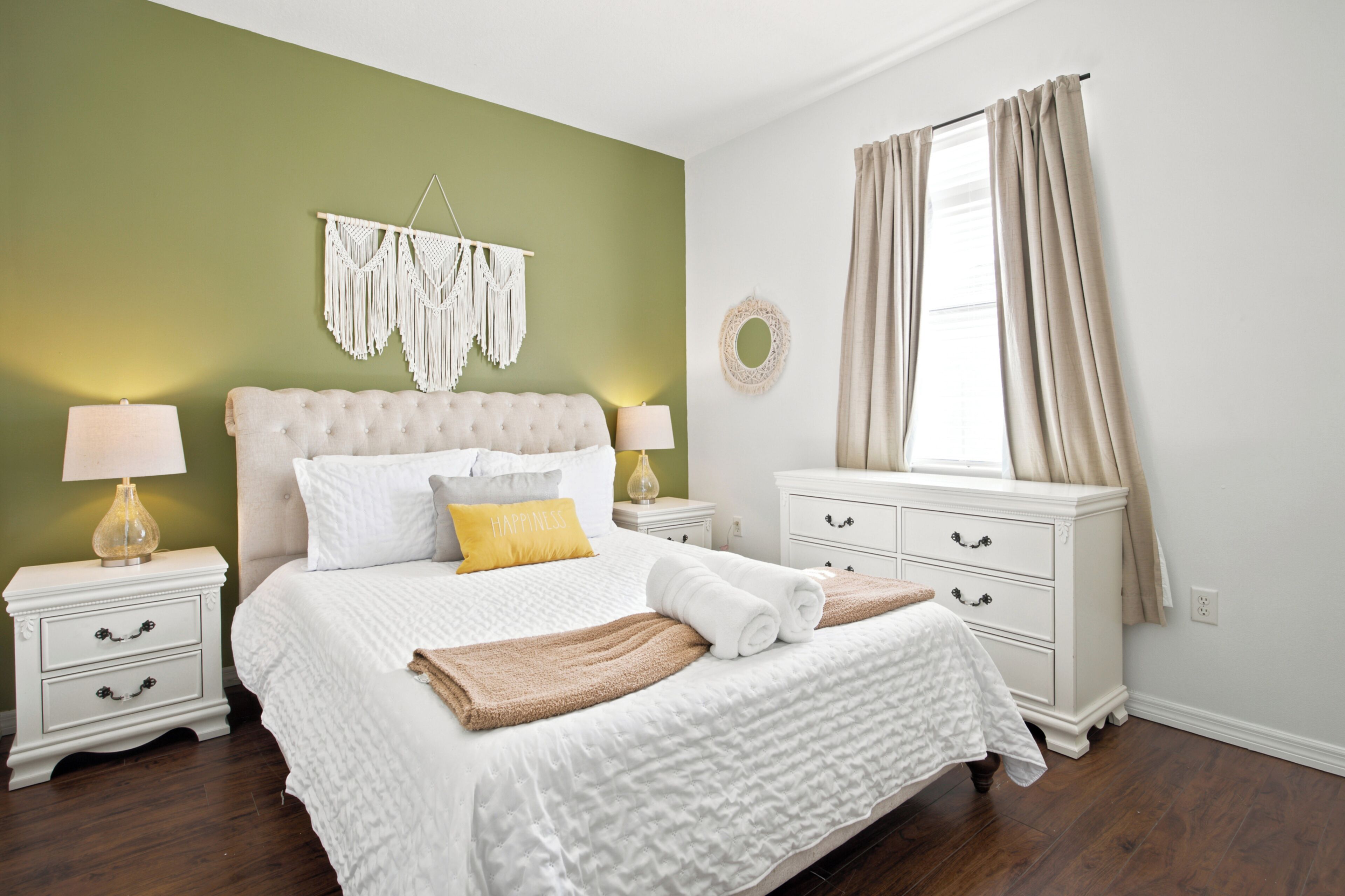 A bedroom with a bed, nightstands, a dresser, a window with curtains, and a wall hanging.
