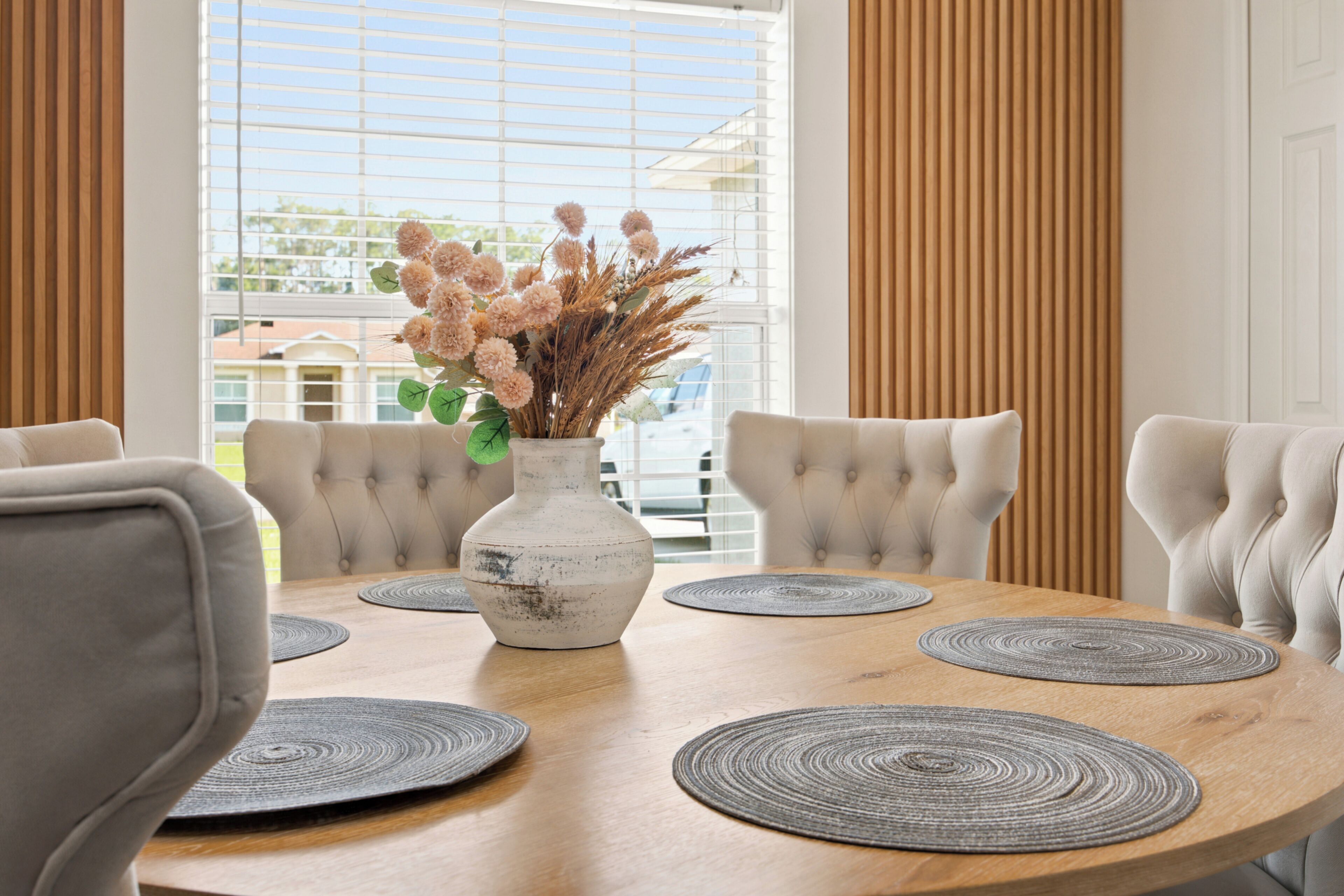 Une table de salle à manger ronde, surmontée d’un vase blanc et agrémentée de fleurs décoratives, entourée de six chaises rembourrées.