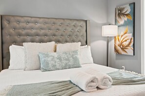 Una cama bien hecha con un cabecero capitonado, varios almohadones y una toalla blanca enrollada.
