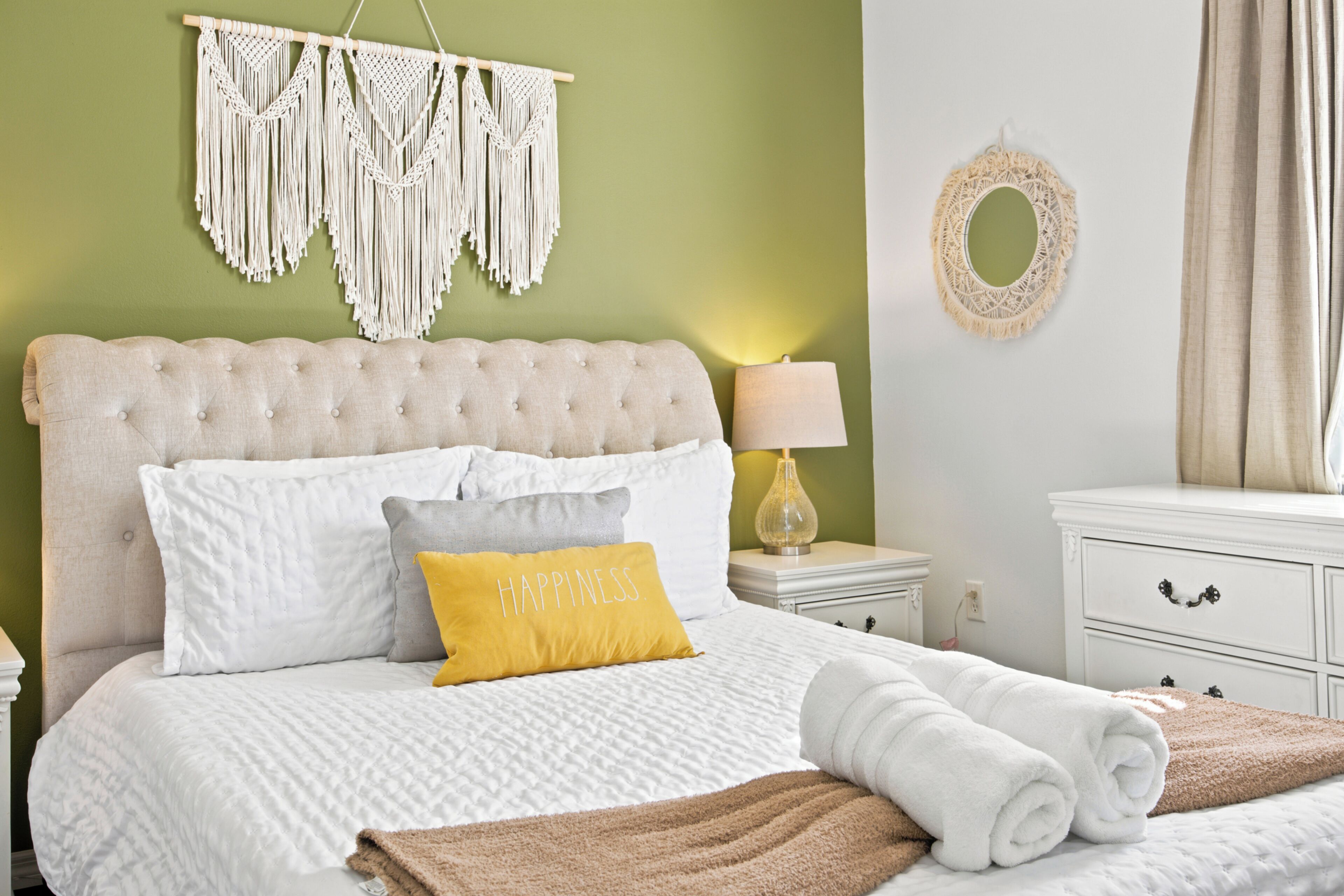 A neatly made bed with white and yellow pillows, a macrame wall hanging, a bedside lamp, and a white dresser.