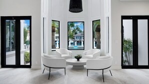 A spacious room with white walls, large windows, and a black ceiling light. There are two white armchairs and a small round table with a plant. - WATERFRONT MANSION (Fort Lauderdale)