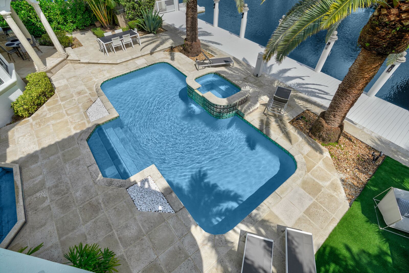 A swimming pool with a small hot tub, surrounded by palm trees and a patio area with outdoor furniture.