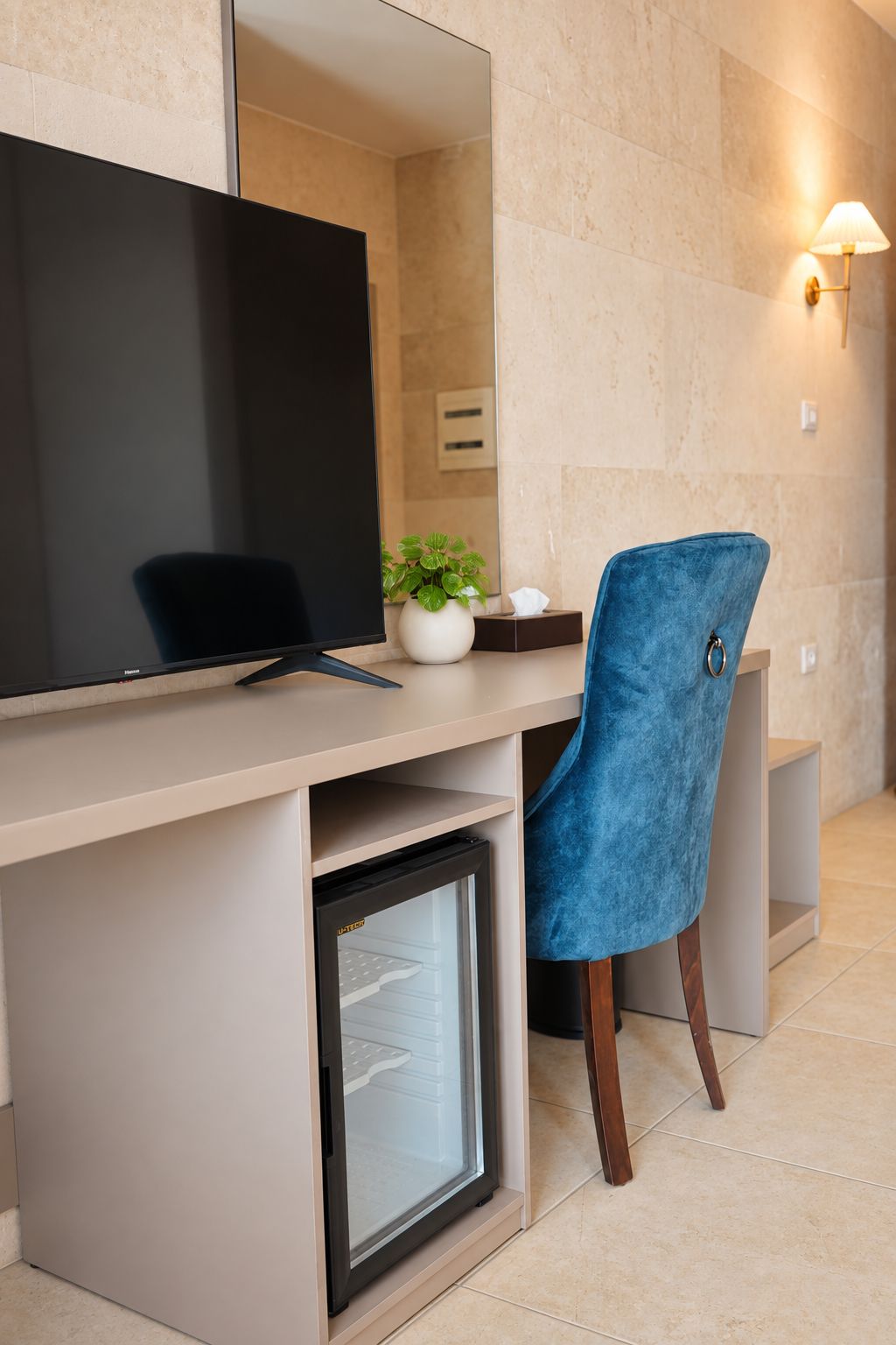 Een moderne kantoorruimte met een bureau, een computerscherm, een kleine koelkast, een blauwe stoel, een potplant en een wandlamp.