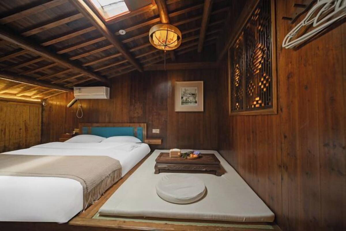 Una habitación de madera con una cama, una mesita con un libro y fruta, y un cuadro en la pared.