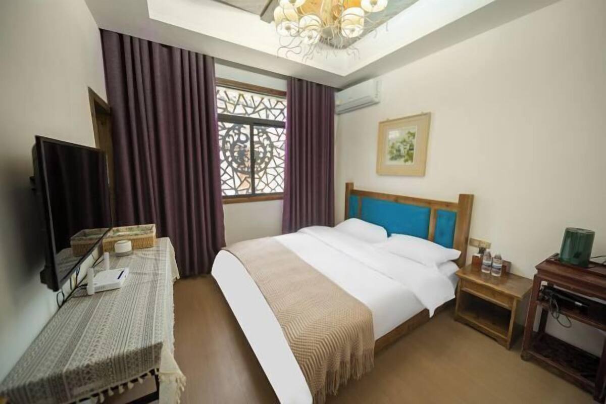 Un cuarto de hotel con cama, escritorio, silla, televisor, ventana con cortinas, un cuadro en la pared y una lámpara de araña.