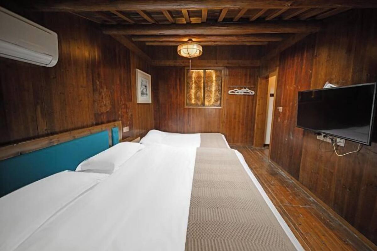 Una habitación con paredes de madera, ventilador de techo, televisor y dos camas con sábanas blancas.
