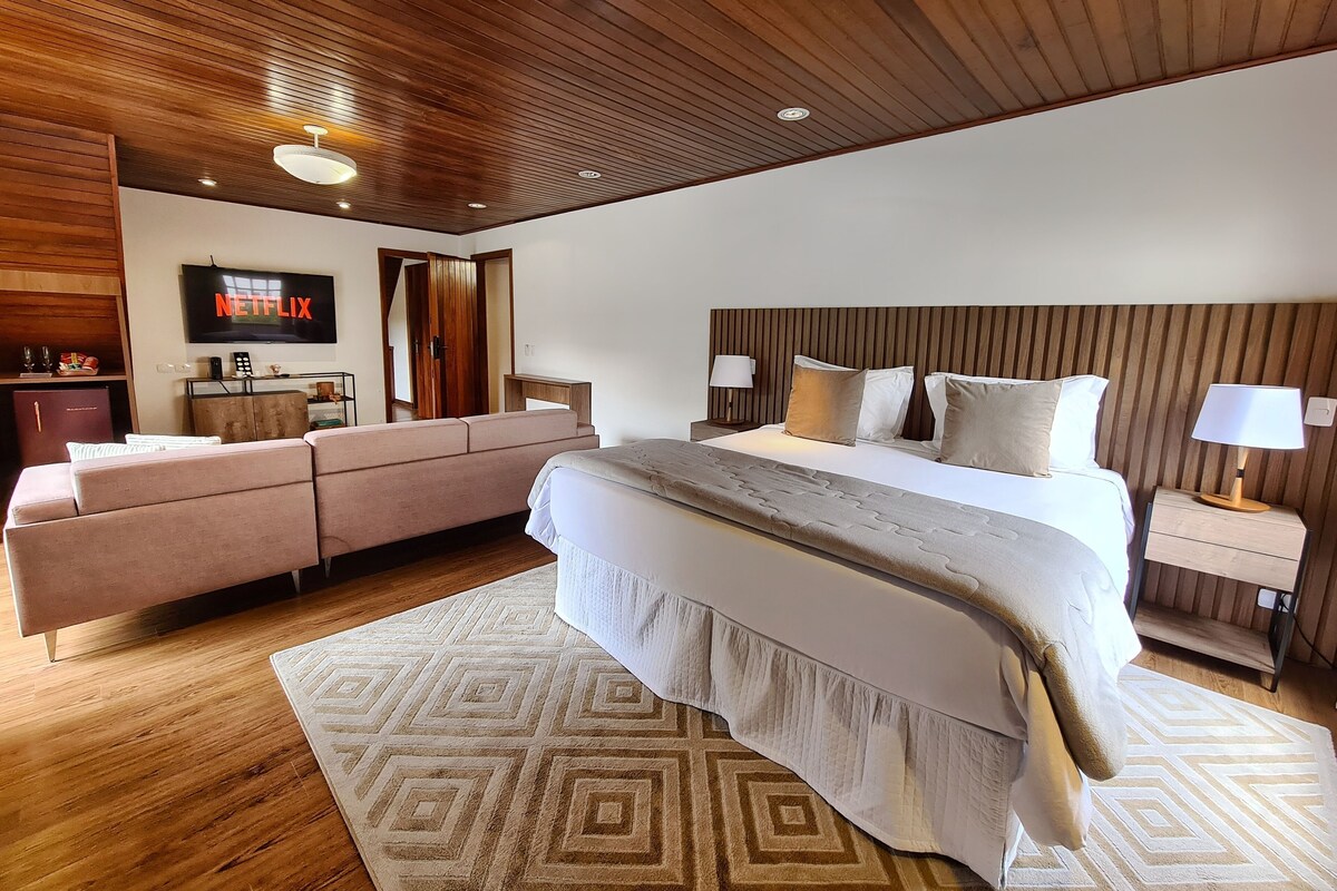 Un dormitorio moderno con techo de madera, una cama grande con cabecera, un sofá, un televisor montado en la pared y una mesita de noche de madera con lámpara.
