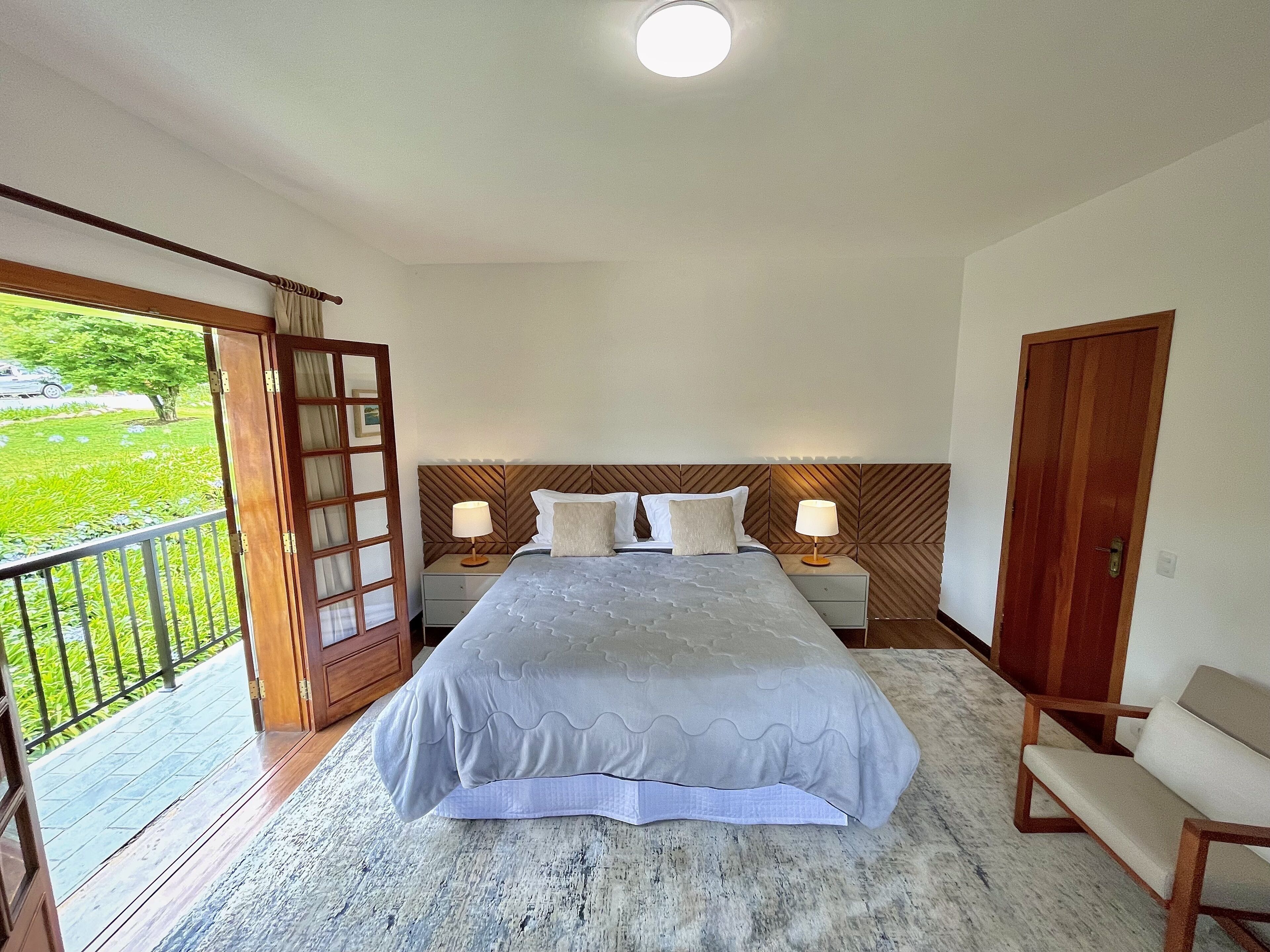 A bedroom with a bed, two bedside tables with lamps, a wooden headboard, a wooden door, and a balcony with a view of greenery.