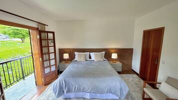 A bedroom with a bed, two bedside tables with lamps, a wooden headboard, a wooden door, and a balcony with a view of greenery.