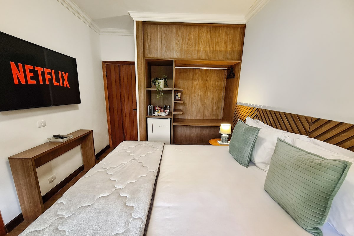 A hotel room with a bed, a TV mounted on the wall, a wardrobe, and a small desk with a lamp.