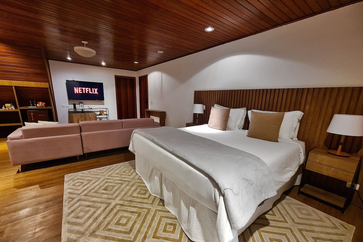 A modern hotel room with a large bed, a sofa, a TV displaying Netflix, wooden ceiling, and a patterned rug.
