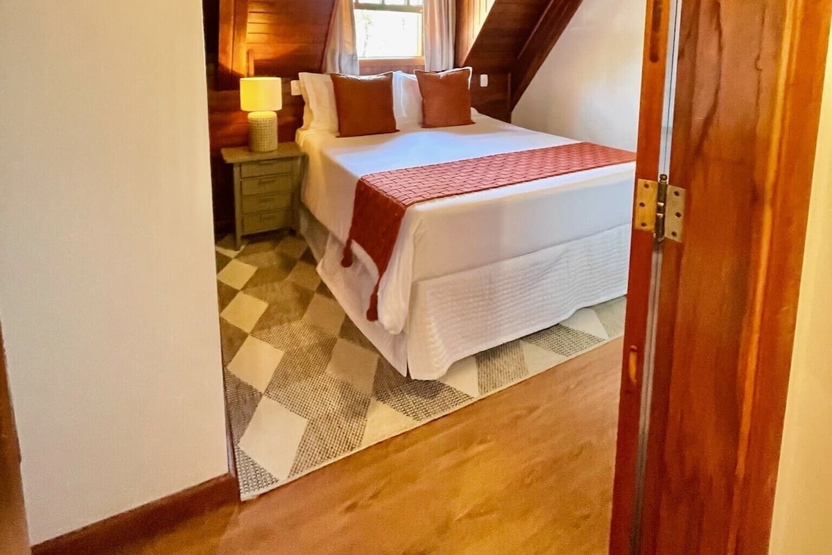 A bedroom with a checkered floor, a bed with a red blanket, wooden ceiling beams, a nightstand, and a painting on the wall.