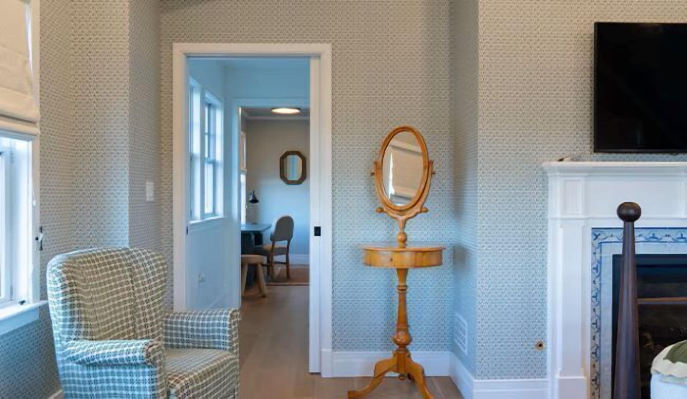 A room with a checkered armchair, a mirror on a pedestal, a fireplace, and a hallway leading to another room.