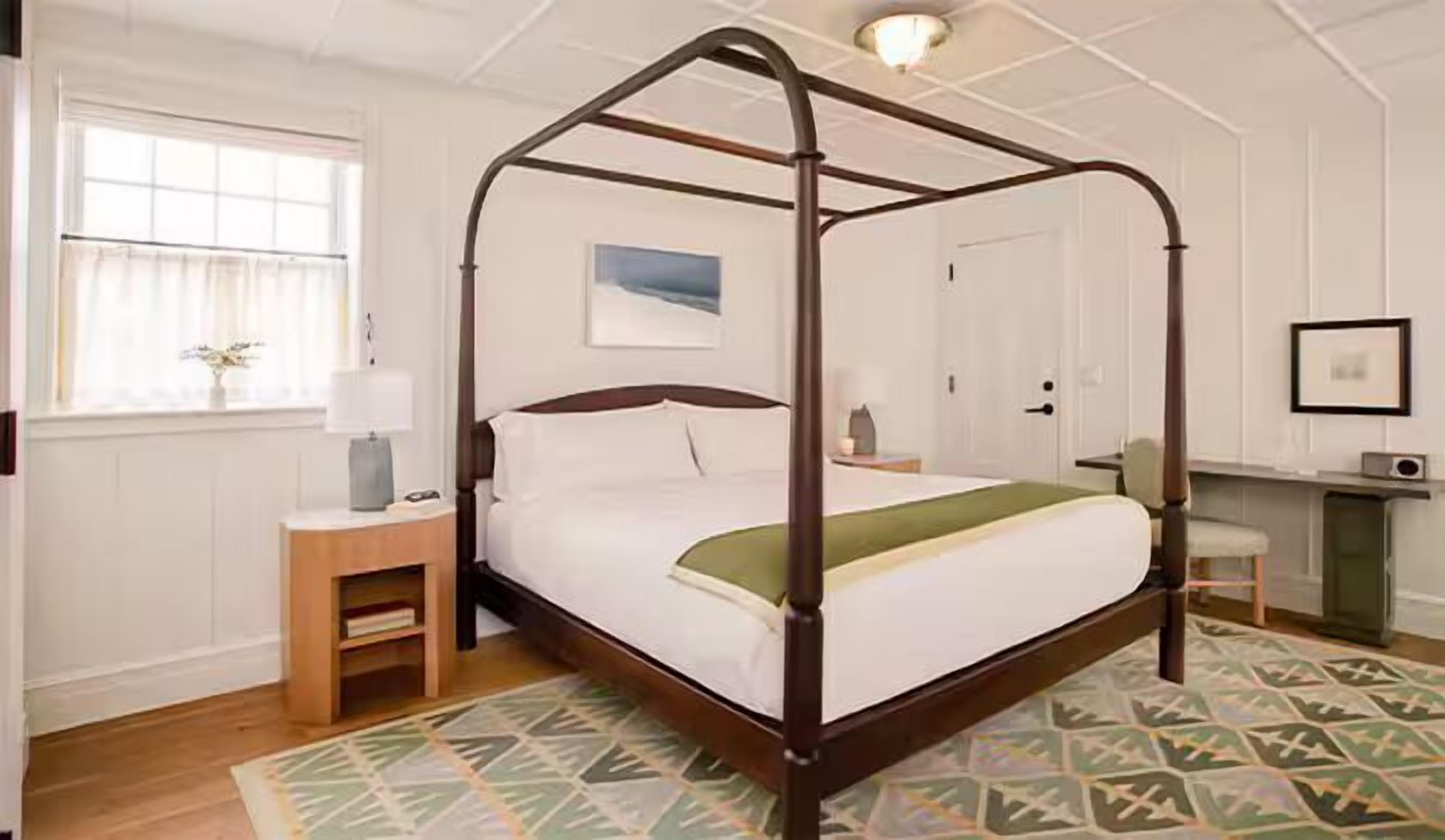 A four-poster bed with a wooden frame, a bedside table with a lamp, a desk with a chair, and a patterned rug.