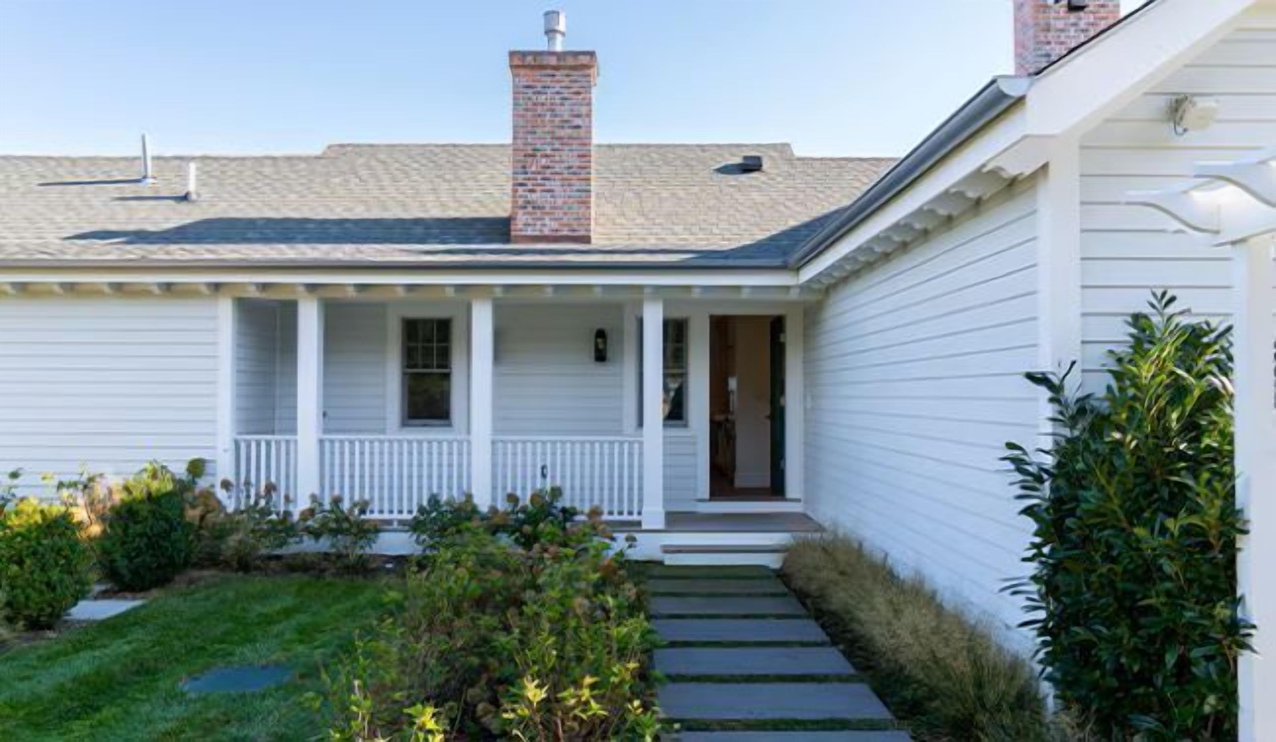 A white single-story house with a front porch, a brick chimney, and a small garden with a pathway leading to the door.