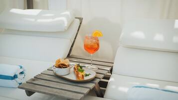 Uma espreguiçadeira com uma mesa na frente, um copo de bebida com uma fatia de laranja e um prato de comida.