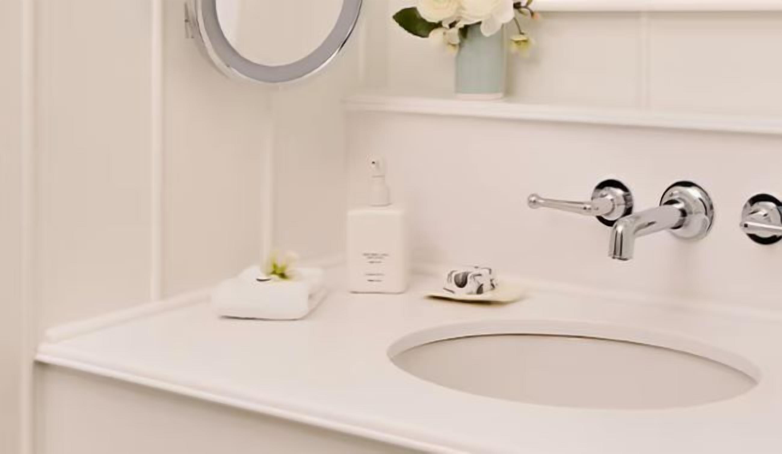 A bathroom with a white sink, a mirror, a faucet, and a small white vase with flowers.