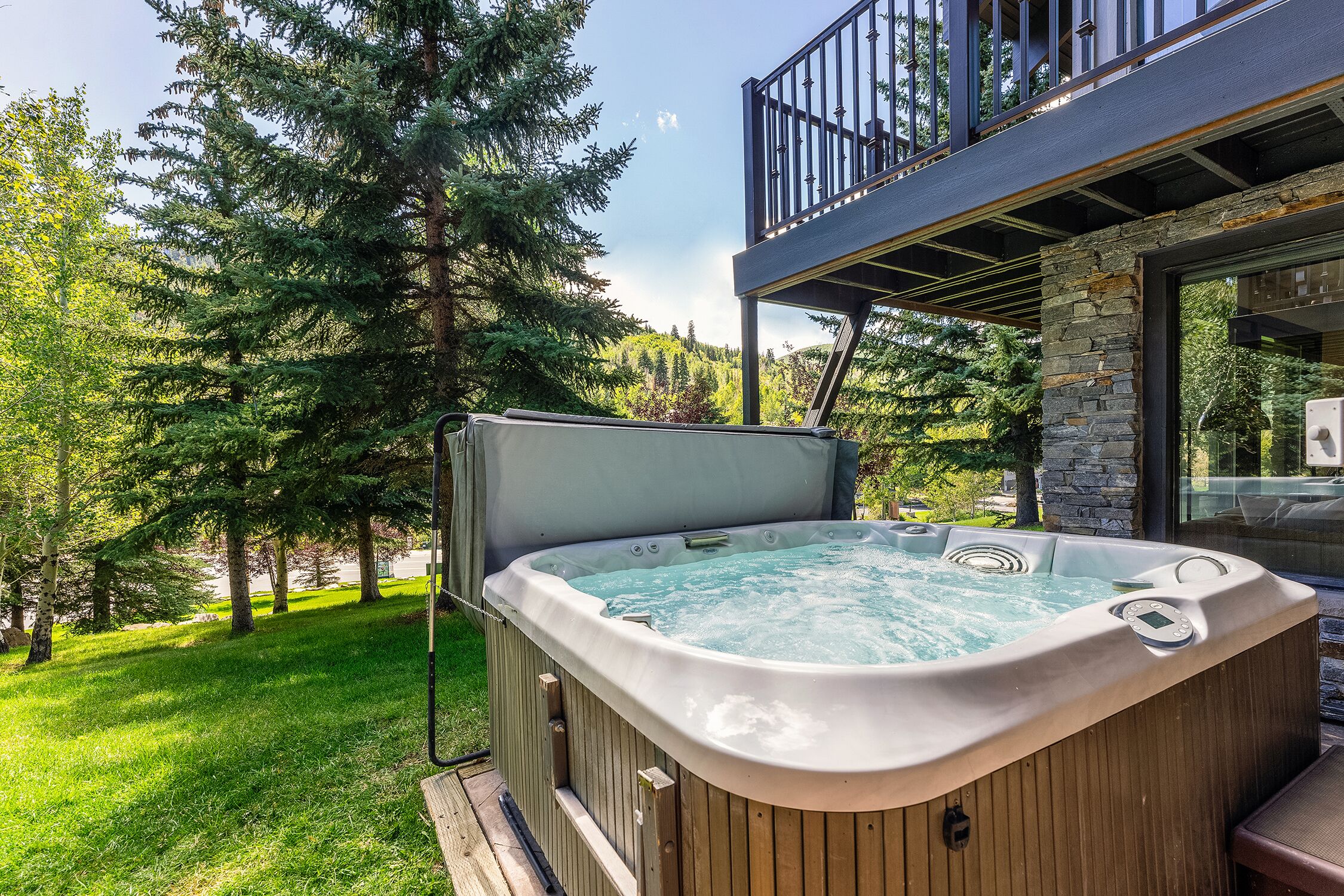 Un jacuzzi en una terraza de madera, con una pared de piedra y puertas de cristal, rodeado de árboles.