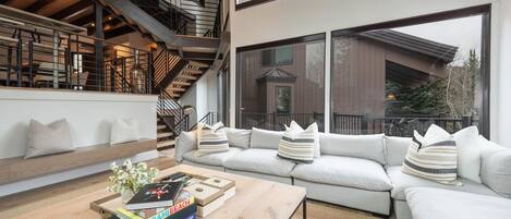 Un salón moderno con sofá, mesa de centro y libros. Se ve una escalera con barandilla al fondo.
