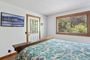 A bedroom with a bed, a wooden bench, a door, a window, and a wall-mounted TV.