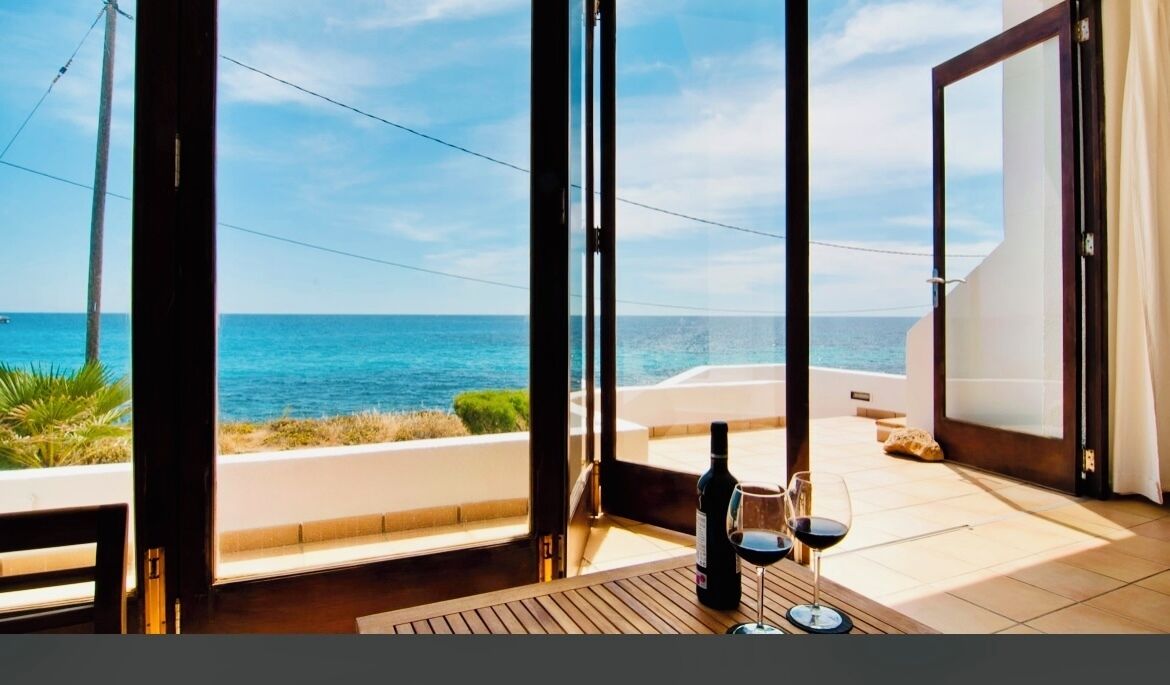 A balcony with a view of the sea, a bottle of wine, and a glass on a wooden table.
