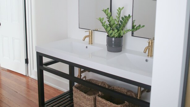 A bathroom with a white vanity, two gold faucets, a black shelf with baskets, and a potted plant. - Brand New 4bd, 2ba Lancaster Center City Townhome (Lancaster)
