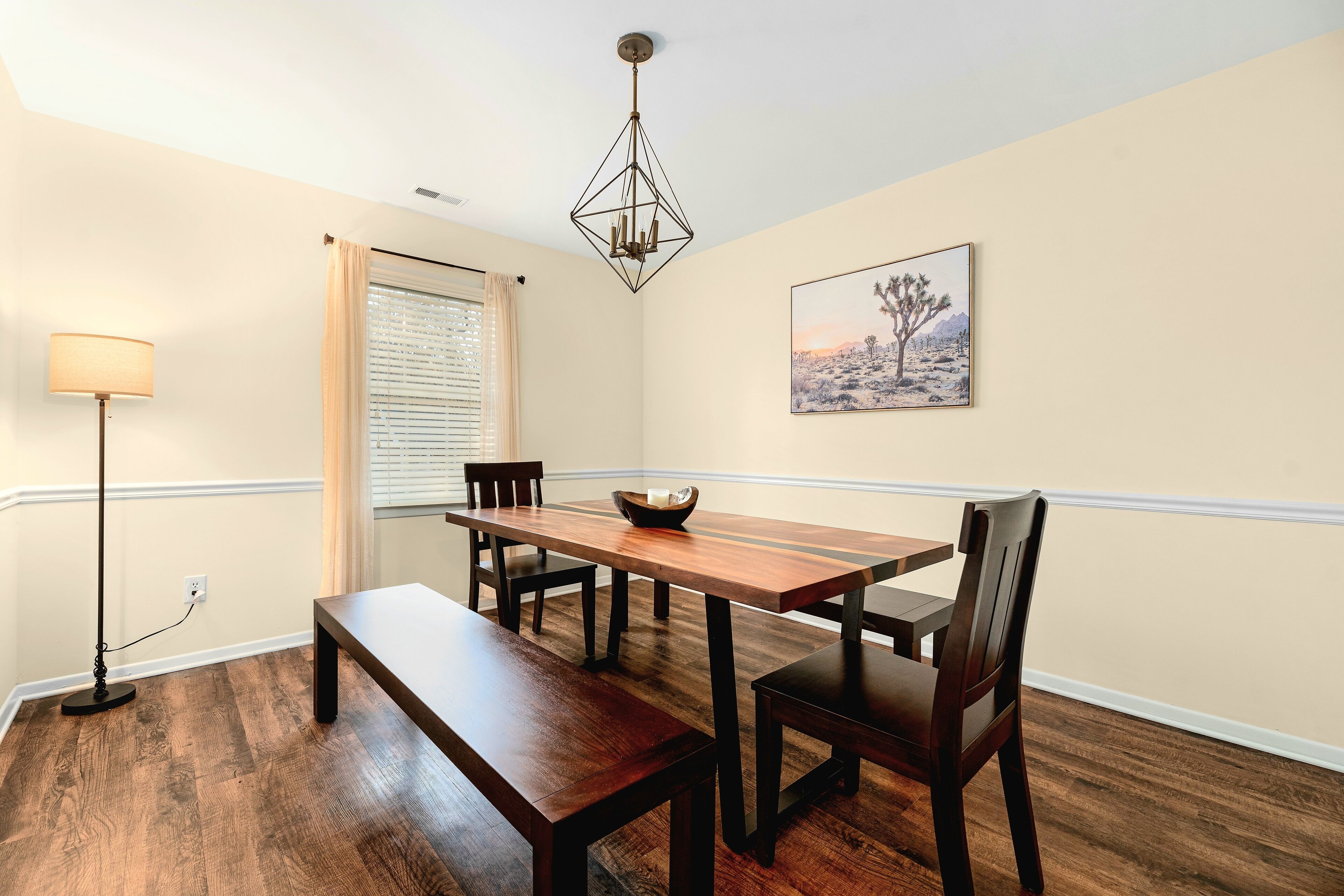 A dining area with a wooden table, benches, and chairs. There is a floor lamp, a window with blinds, and a painting on the wall.
