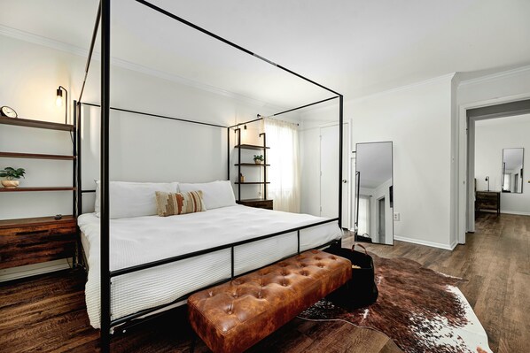 A four-poster bed with a wooden bench, white bedding, and a small plant on a shelf.
