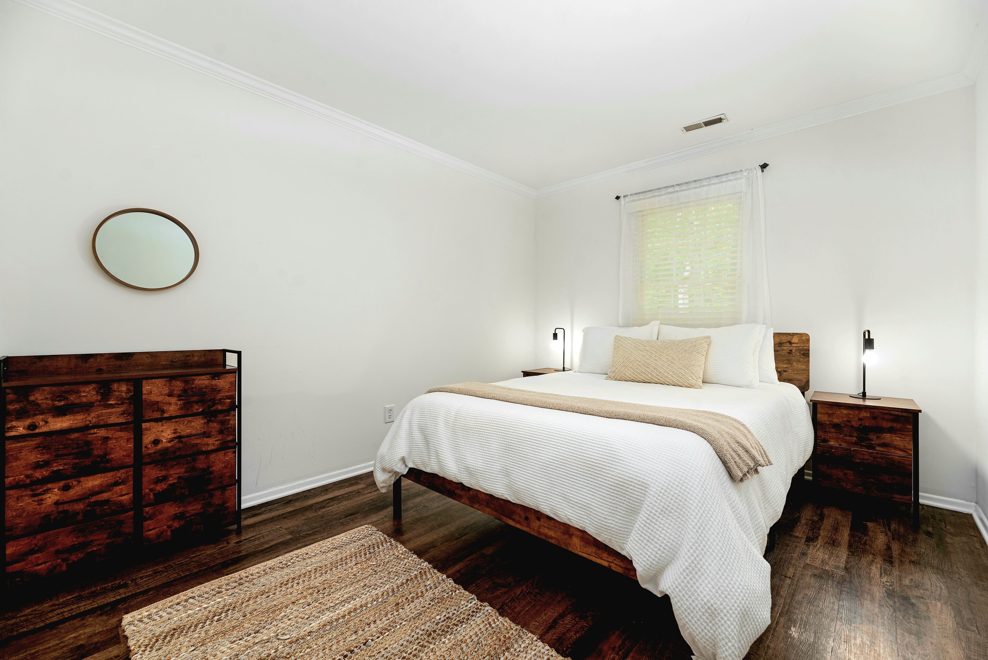 A bedroom with a wooden bed frame, white bedding, a round mirror on the wall, and a small bedside table with a lamp.