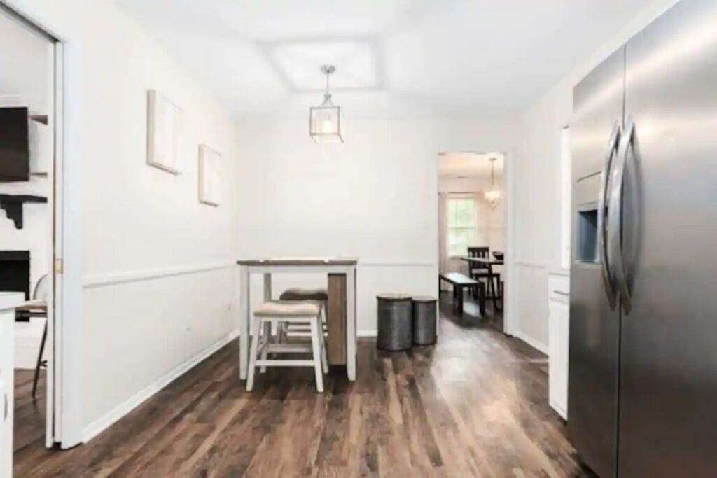 A narrow hallway with a wooden floor, a white wall, a small table with a chair, a trash bin, and a refrigerator.