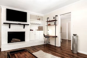 A living room with a white brick fireplace, a flat-screen TV mounted above it, and a wooden coffee table. There are shelves with decorative items and a tall, narrow vase on the wall.