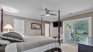 A bedroom with a four-poster bed, a ceiling fan, a TV, and a sliding glass door to a backyard. - Walk to Beach • Hot Tub • Pool Table • Ideal for Long Stays (Oak Island)