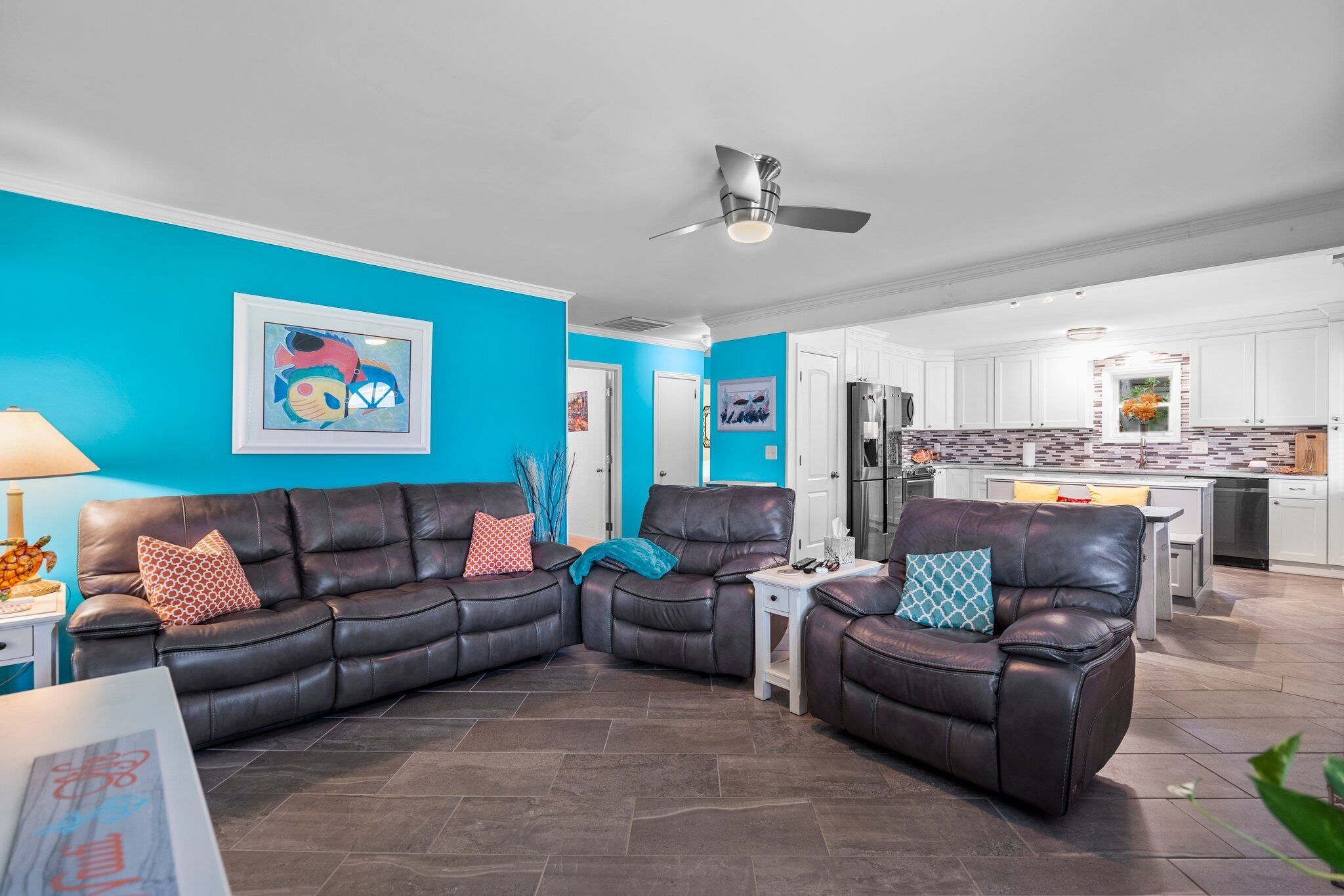Un salon avec un canapé d’angle, un ventilateur de plafond et une cuisine en arrière-plan.