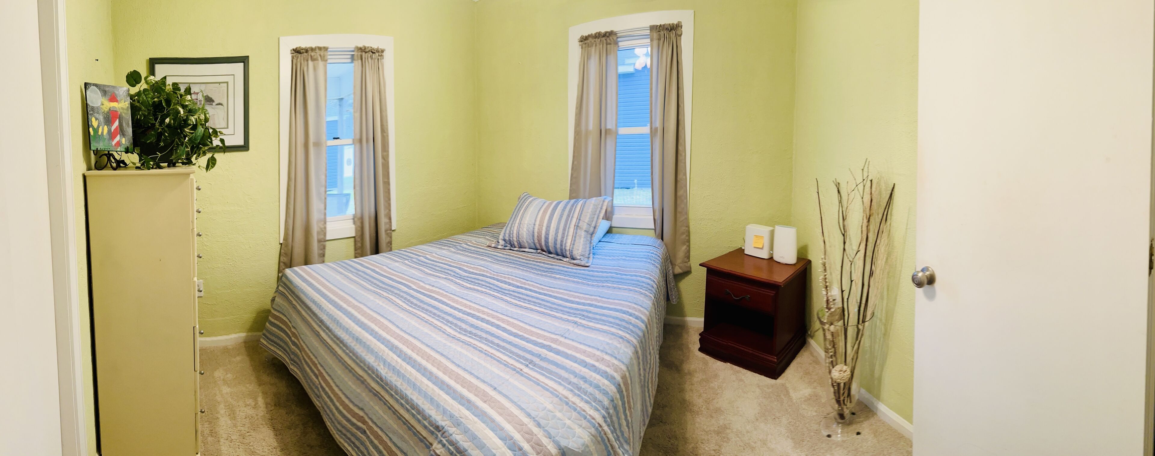 A bedroom with a bed, two windows with curtains, a nightstand, a dresser, a plant, and wall decorations.