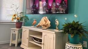 A living room with teal walls, a white entertainment center, a flat-screen TV mounted on the wall, and a potted plant on the side table. - Walk to Beach • Hot Tub • Pool Table • Ideal for Long Stays (Oak Island)