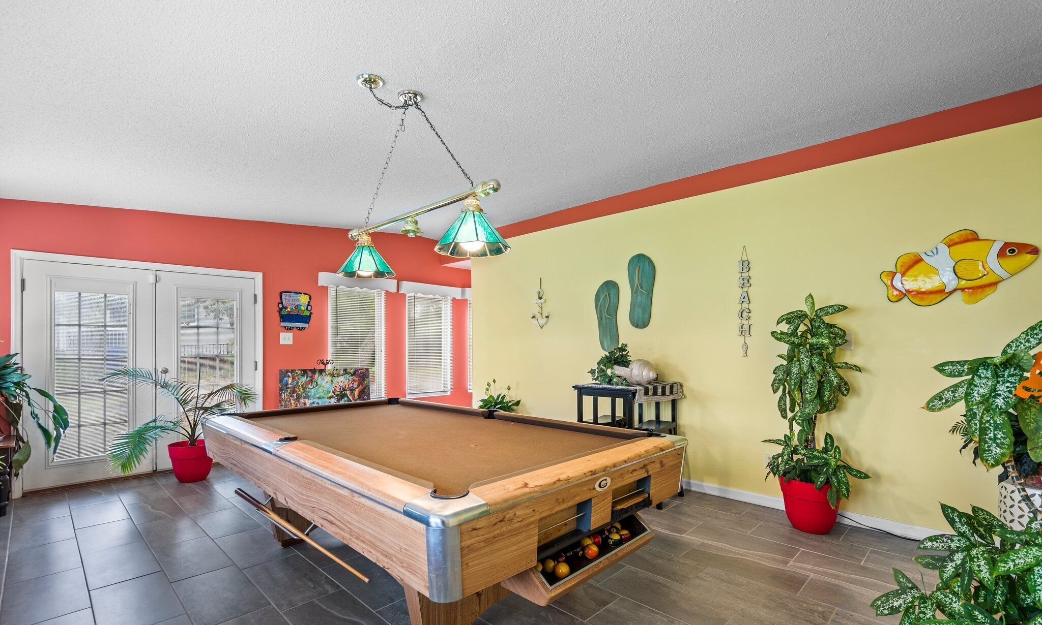 Une table de billard dans une pièce aux murs rouge et jaune, avec un ventilateur de plafond et plusieurs plantes en pot.