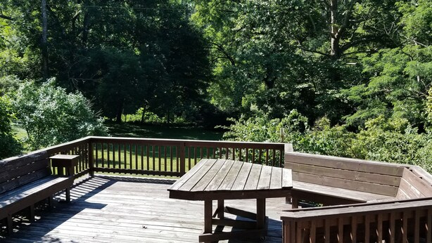 A wooden deck with benches and a table, surrounded by dense forest. - Nestled near woods and creek, this large home is near state parks and colleges.. (Hillsboro)