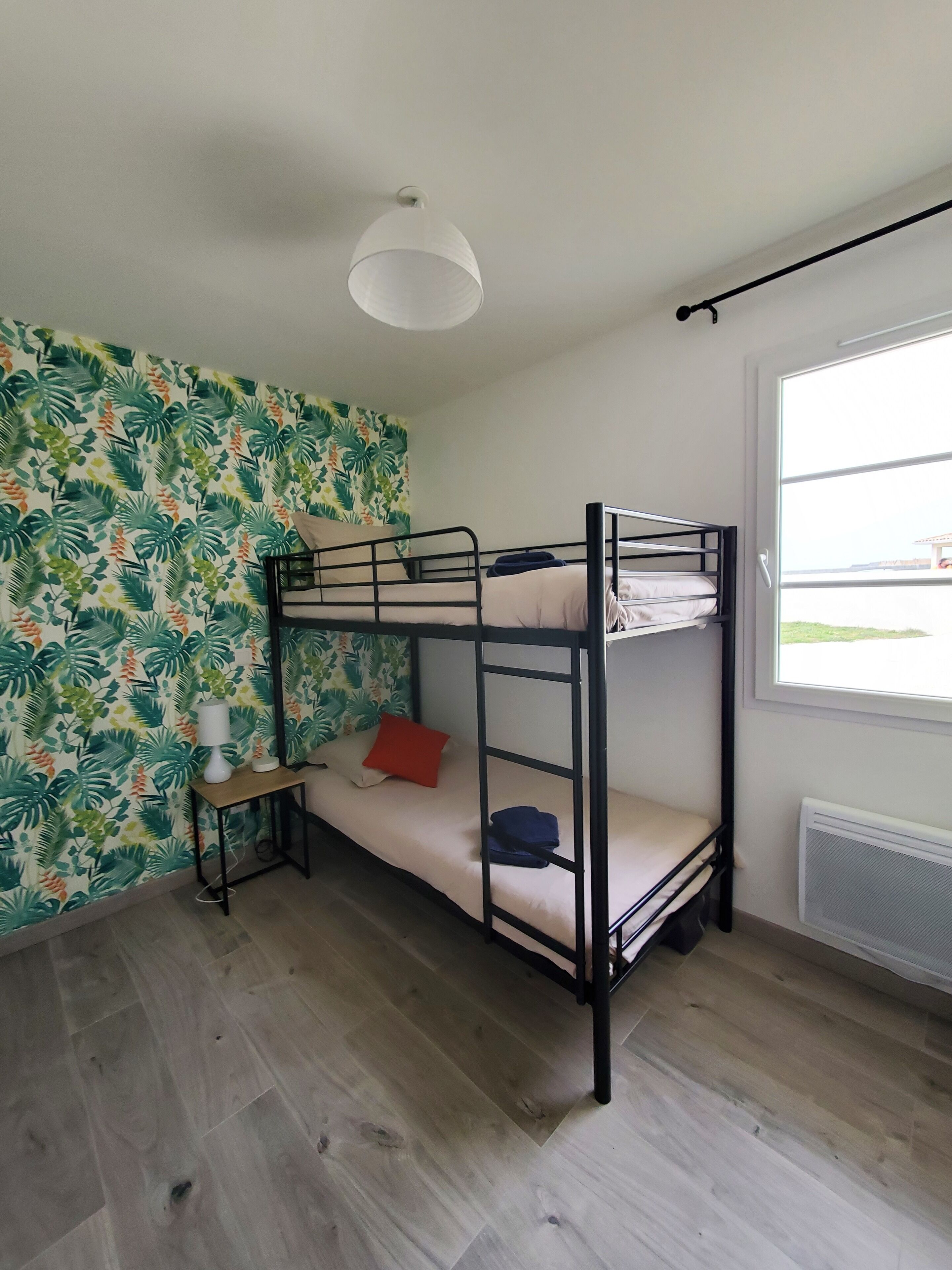 Habitación con literas, papel tapiz tropical, una mesita de noche pequeña con lámpara y ventana con vista al exterior.