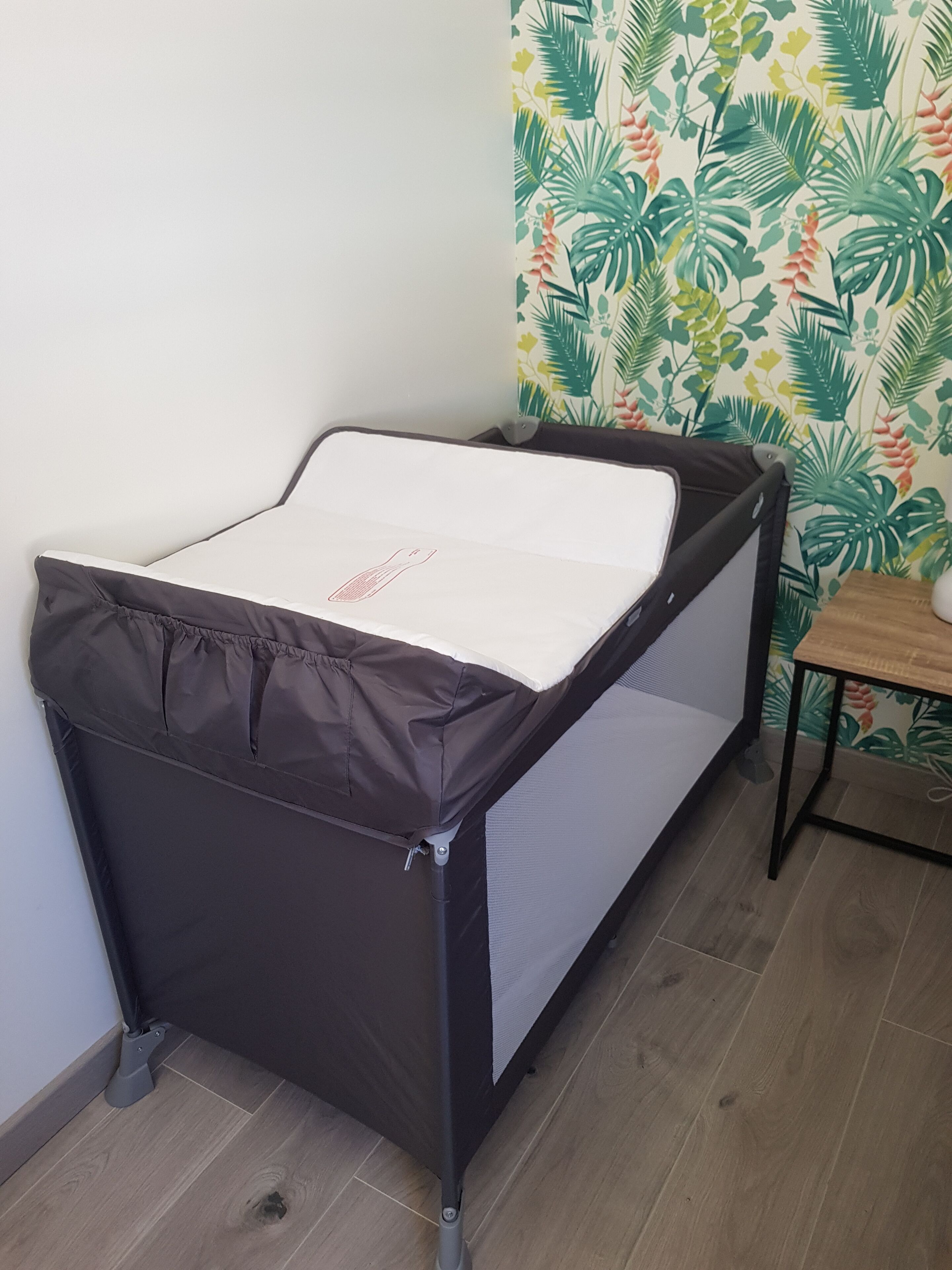 A baby changing table with a protective cover, positioned against a wall with a tropical leaf wallpaper.