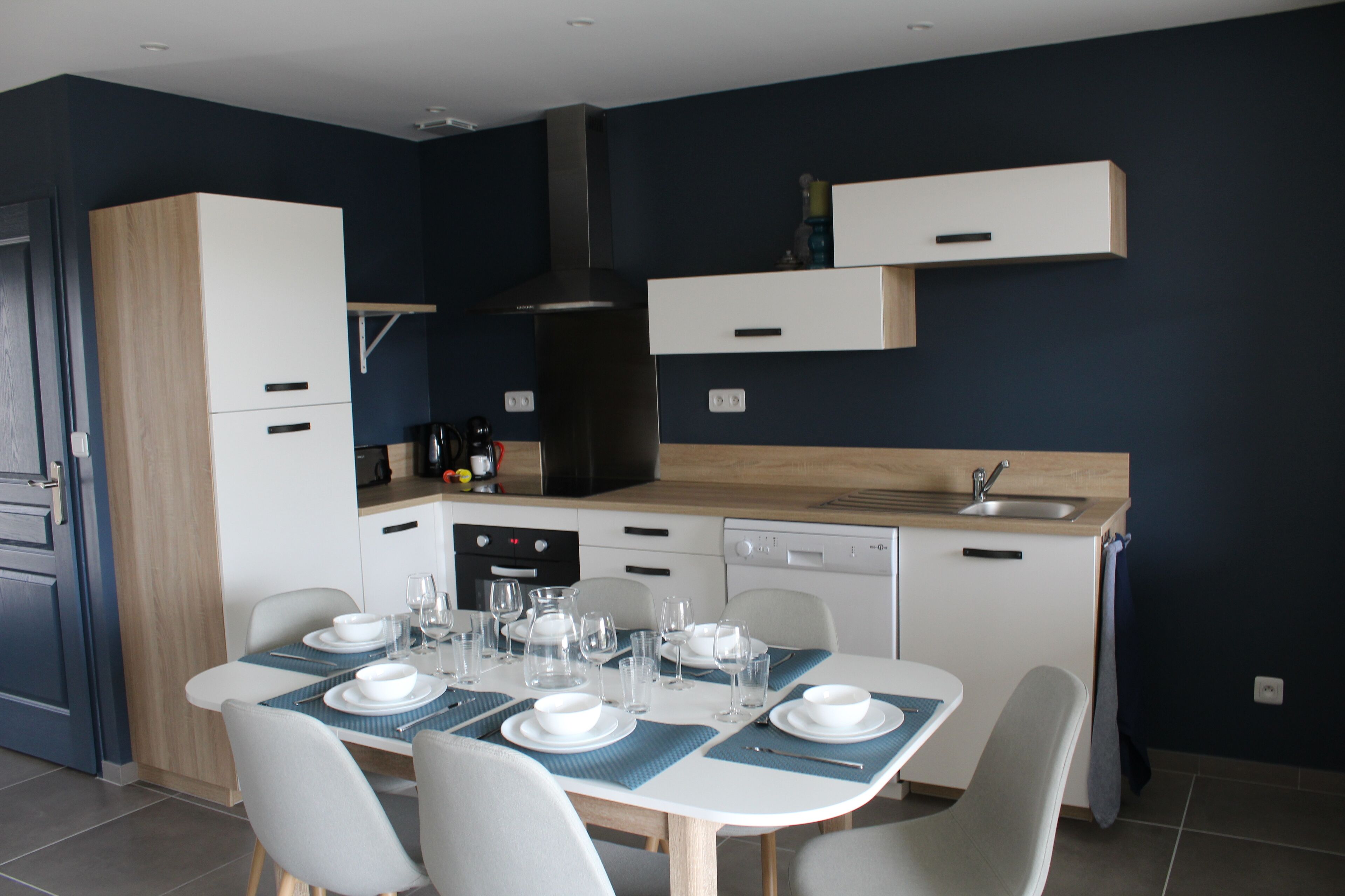 A modern kitchen with white cabinets, a built-in oven, and a dishwasher. There is a dining table set with plates, glasses, and cutlery.