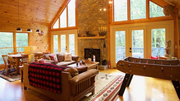 A cozy cabin interior with a stone fireplace, wooden furniture, and large windows. - Luxury Blue Ridge Cabin with Hot Tub & Game Room - Sleeps 8 (Ferguson)