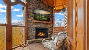 Une terrasse en bois avec un foyer, des fauteuils en osier et un téléviseur fixé à un mur en pierre.