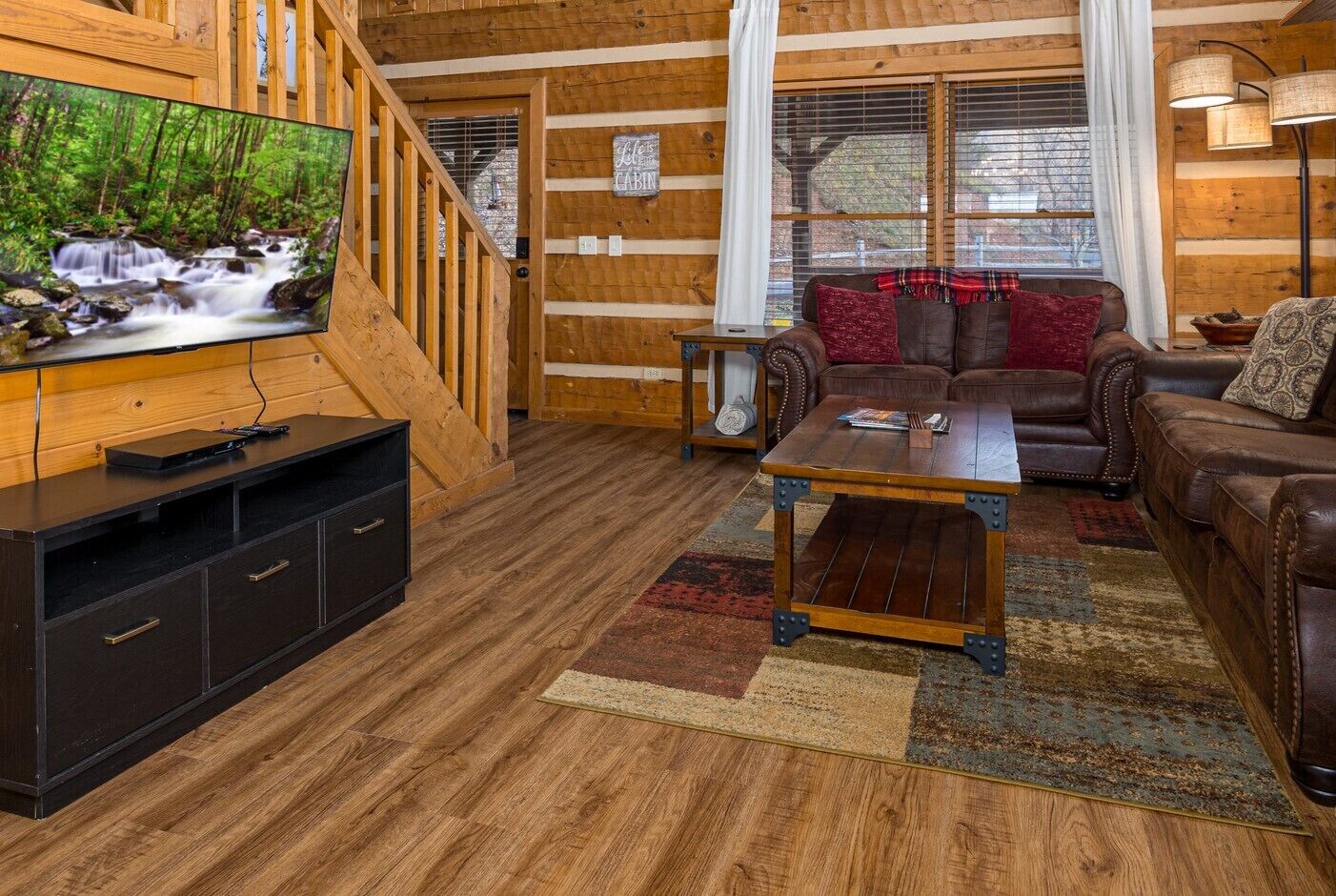 A wooden cabin interior with a TV, leather sofas, a coffee table, and a staircase.