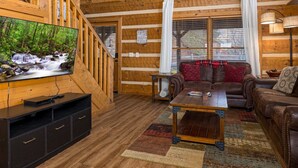 A wooden cabin interior with a TV, leather sofas, a coffee table, and a staircase.