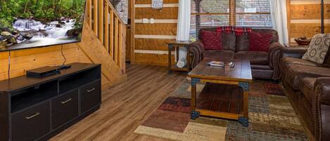 A wooden cabin interior with a TV, leather sofas, a coffee table, and a staircase.